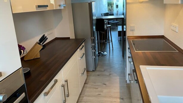 A modern kitchen with white cabinets, a dark countertop, and a central island. There is a dining area with a table and chairs visible through an archway. - Apartment in Alsdorf - near Aachen (Alsdorf)