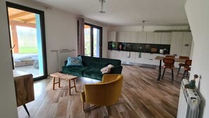 Amplia sala de estar con un sofá verde, un sillón amarillo y una mesa de centro de madera. Se observa una cocina con armarios blancos y un comedor con sillas naranjas.