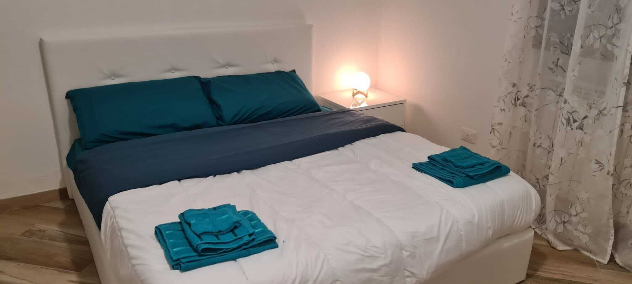 Un lit bien fait, agrémenté de coussins turquoise et de serviettes pliées, une lampe de chevet et un rideau à motifs floraux.