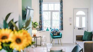 Uma sala de jantar com uma mesa posta para dois, uma janela com cortinas, uma televisão fixada na parede e uma porta com uma placa.