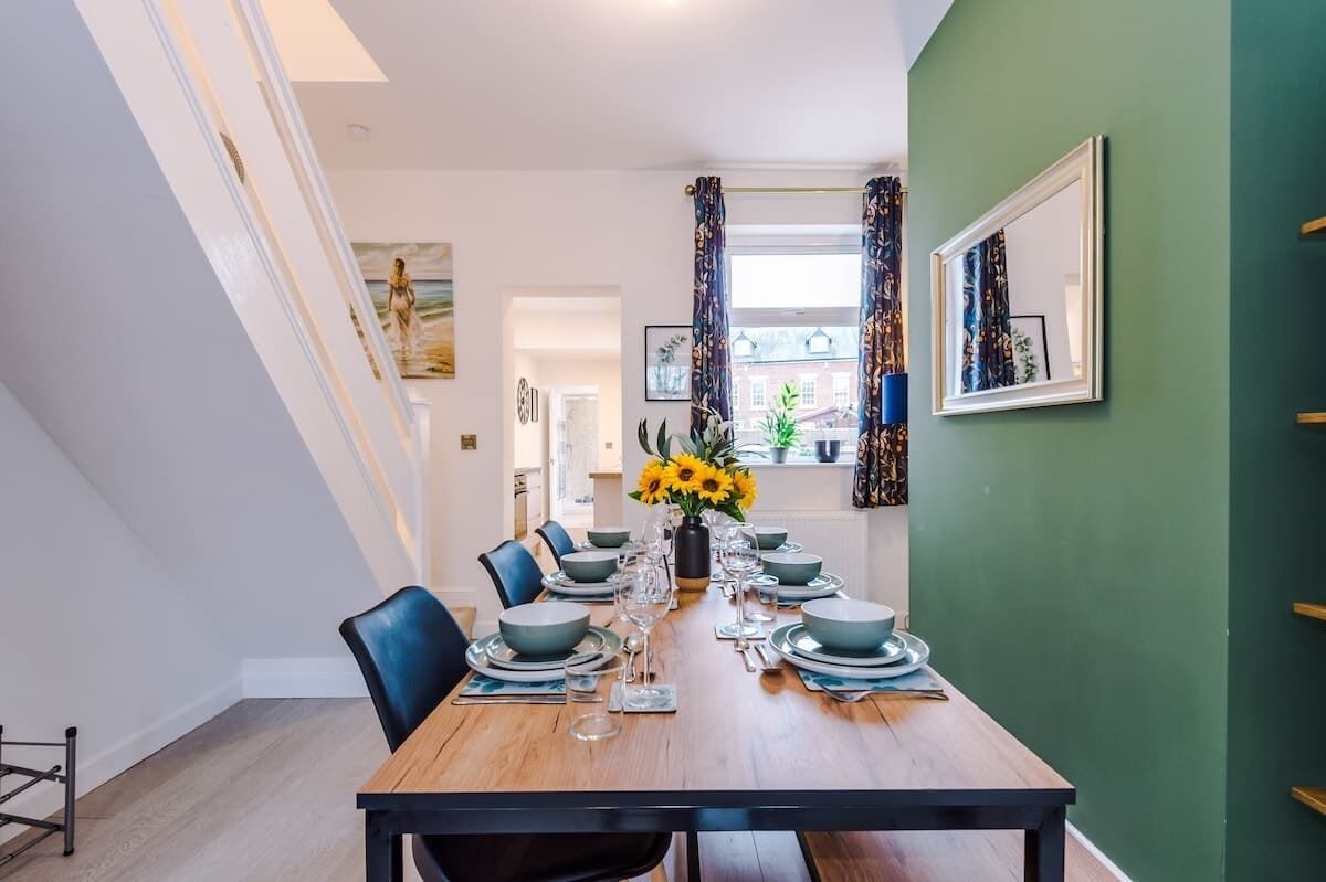 A dining room with a wooden table set for six, surrounded by chairs, with a vase of sunflowers on the table.