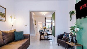 Uma sala de estar moderna com um sofá de couro marrom, uma área de jantar com mesa e cadeiras, e uma parede verde com um painel de TV exibindo 'NETFLIX'.
