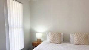 Una habitación de hotel con cama, almohadas, lámpara de noche y ventana con persianas.
