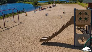 Una playa arenosa con un parque infantil, que incluye columpios y un tobogán, con vistas a un lago y montañas al fondo.