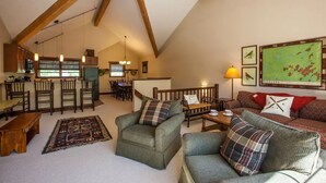 Un espacio habitable tipo loft, acogedor, con techos altos, vigas de madera a la vista, una zona de comedor con una mesa larga y sillas, una sala de estar con sofá y sillones, y una cocina al fondo.