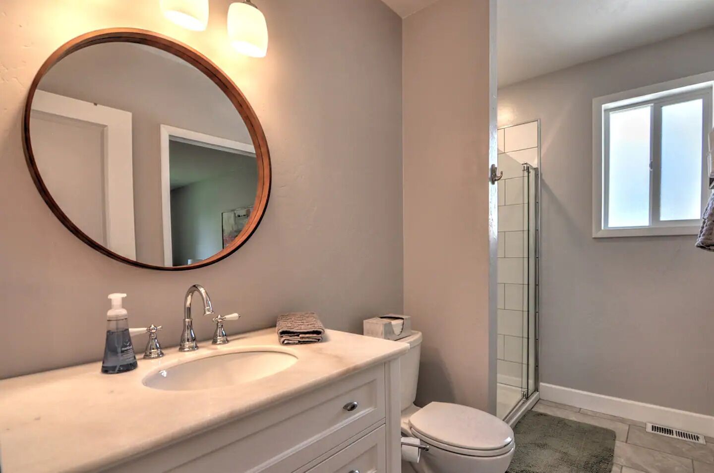 A bathroom with a round mirror, a sink with two faucets, a toilet, and a glass shower enclosure.