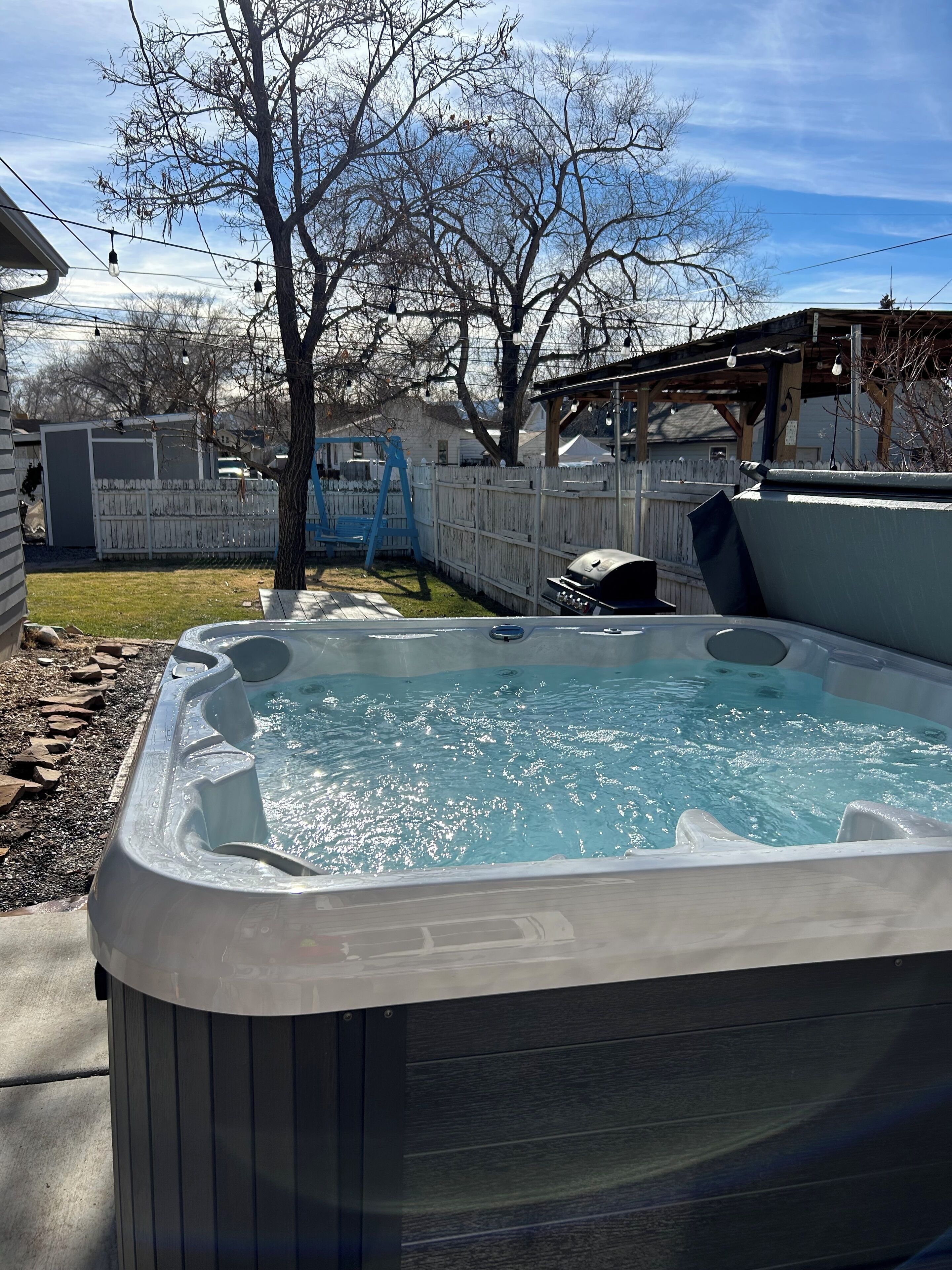 A hot tub in a backyard with a wooden deck, a swing set, and a barbecue grill.