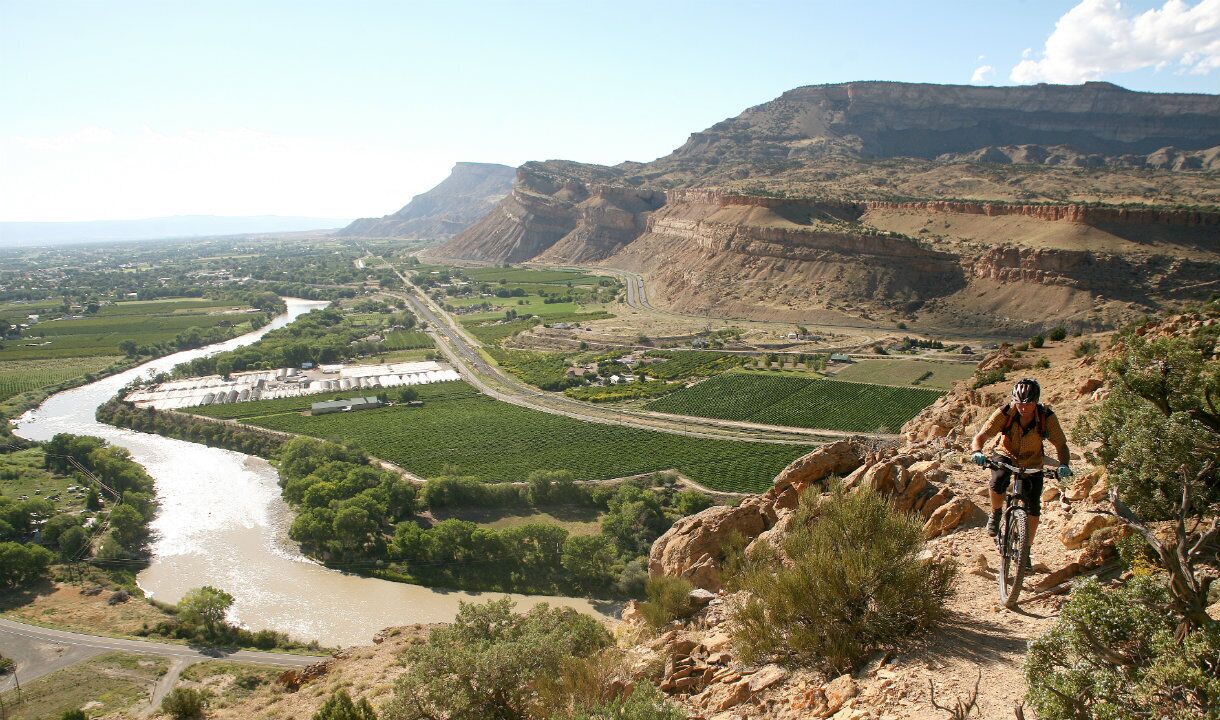 A mountainous landscape with a river, a cyclist, and a bridge.