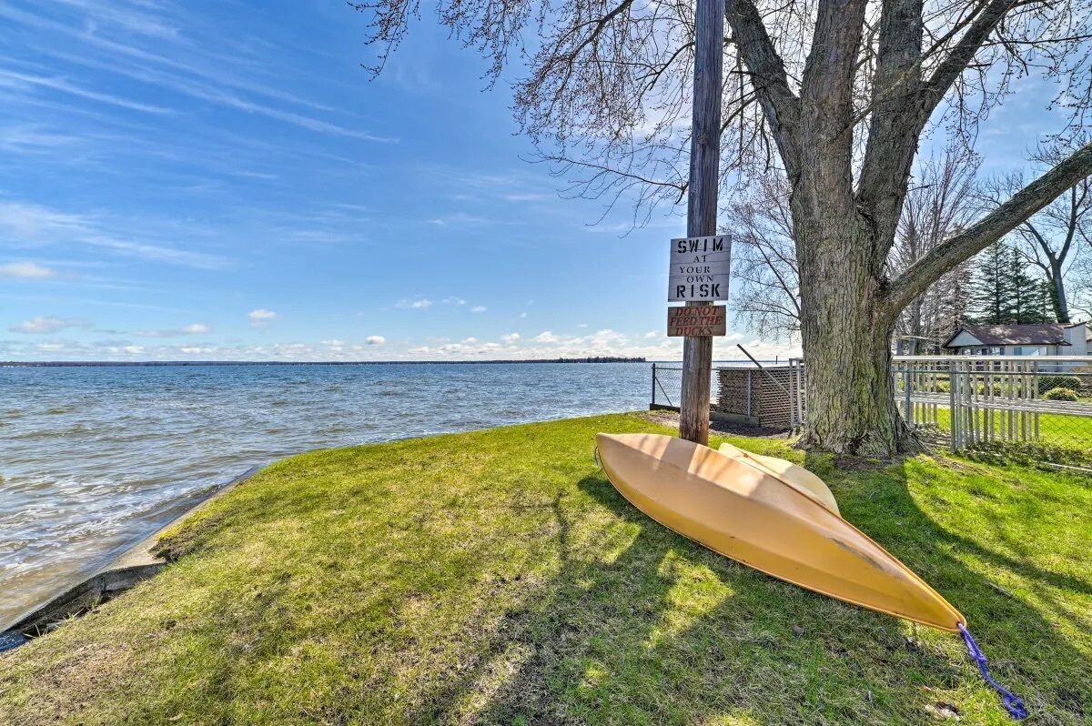 Un kayak est attaché à un arbre au bord d’un lac, et une pancarte indique les risques liés à la baignade et rappelle qu’il est interdit de nourrir les canards.