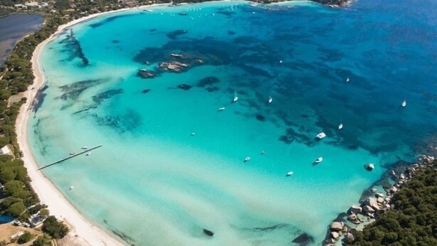 Vue aérienne d’une zone côtière avec une vaste baie aux eaux turquoise, des bateaux et une plage.