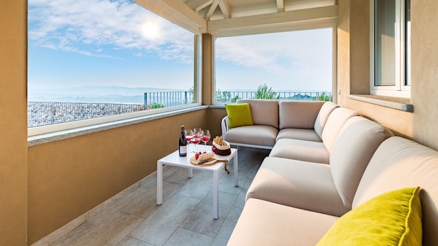 Ein Balkon mit einem beigen Sofa, einem kleinen Tisch mit Weinflaschen und Snacks und Blick aufs Meer.
