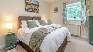 Een netjes opgemaakt bed met witte lakens en een groene gebreide plaid, een bedlampje en een raam met gordijnen.