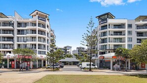 A modern apartment complex with multiple balconies, a central courtyard, and surrounding commercial establishments. - Beachside 2-Bed with Pools, Gym, BBQ and Parking (Alexandra Headland)