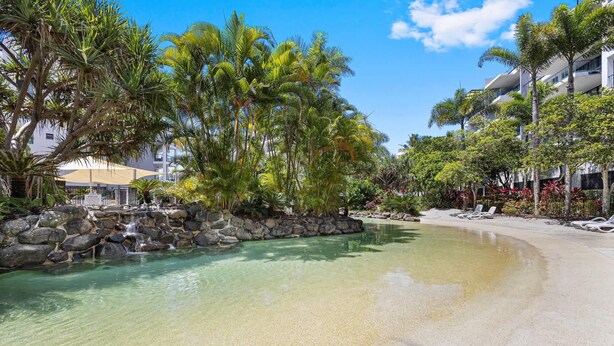 A clear pool with a rocky edge, surrounded by palm trees and a sandy beach. - Beachside 2-Bed with Pools, Gym, BBQ and Parking (Alexandra Headland)