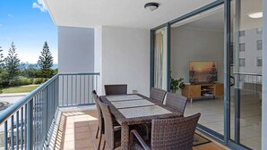 A balcony with wicker furniture, a glass door leading to a room with a painting, and a view of trees and a body of water. - Beachside 2-Bed with Pools, Gym, BBQ and Parking (Alexandra Headland)
