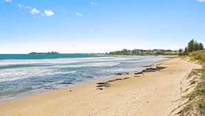 A beach with a clear sky, calm sea, and buildings in the background. - Beachside 2-Bed with Pools, Gym, BBQ and Parking (Alexandra Headland)