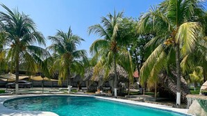 A clear blue pool surrounded by palm trees and thatched-roof structures. - Santorini Village - Hotel Boutique (Tamanique)