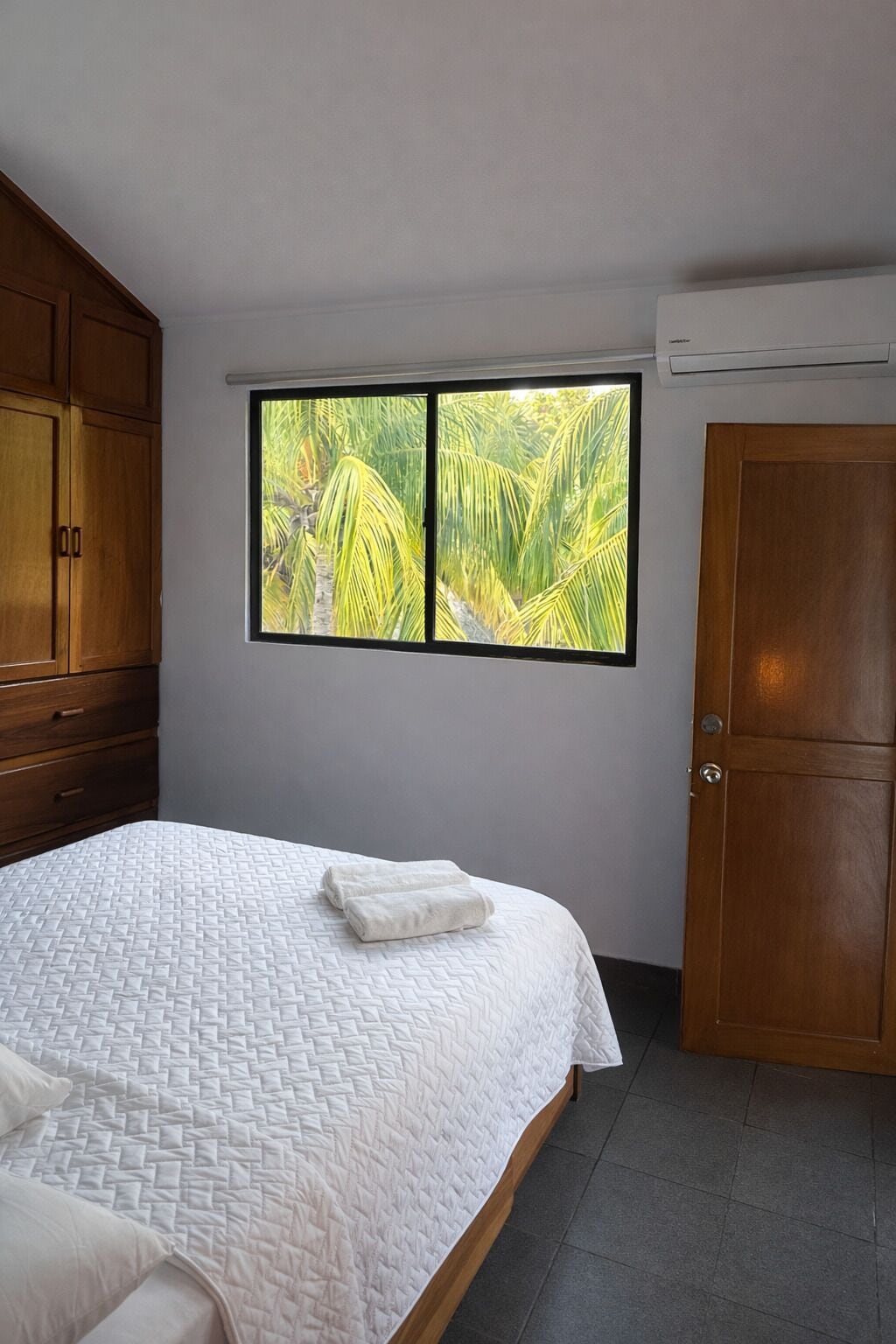 Un dormitorio con cama, una ventana con vistas a palmeras, un armario de madera y una puerta.