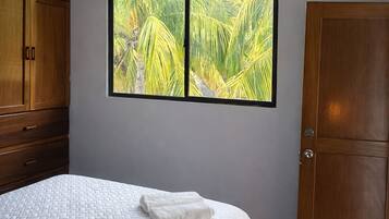 Un dormitorio con cama, una ventana con vistas a palmeras, un armario de madera y una puerta.