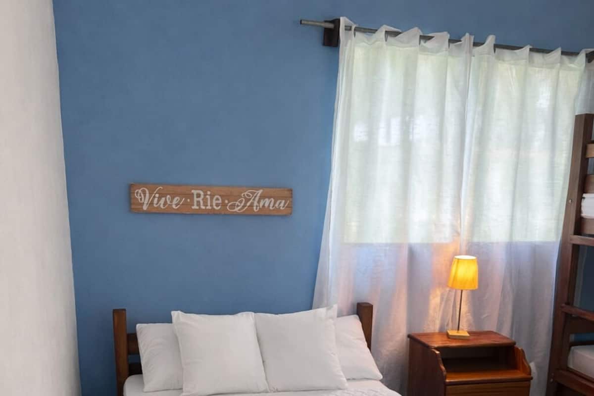 Ein Schlafzimmer mit einem Bett, einem Nachttisch mit Lampe, einem hölzernen Kopfteil, einem an der Wand befestigten Schild und einem Fenster mit durchscheinenden Vorhängen.