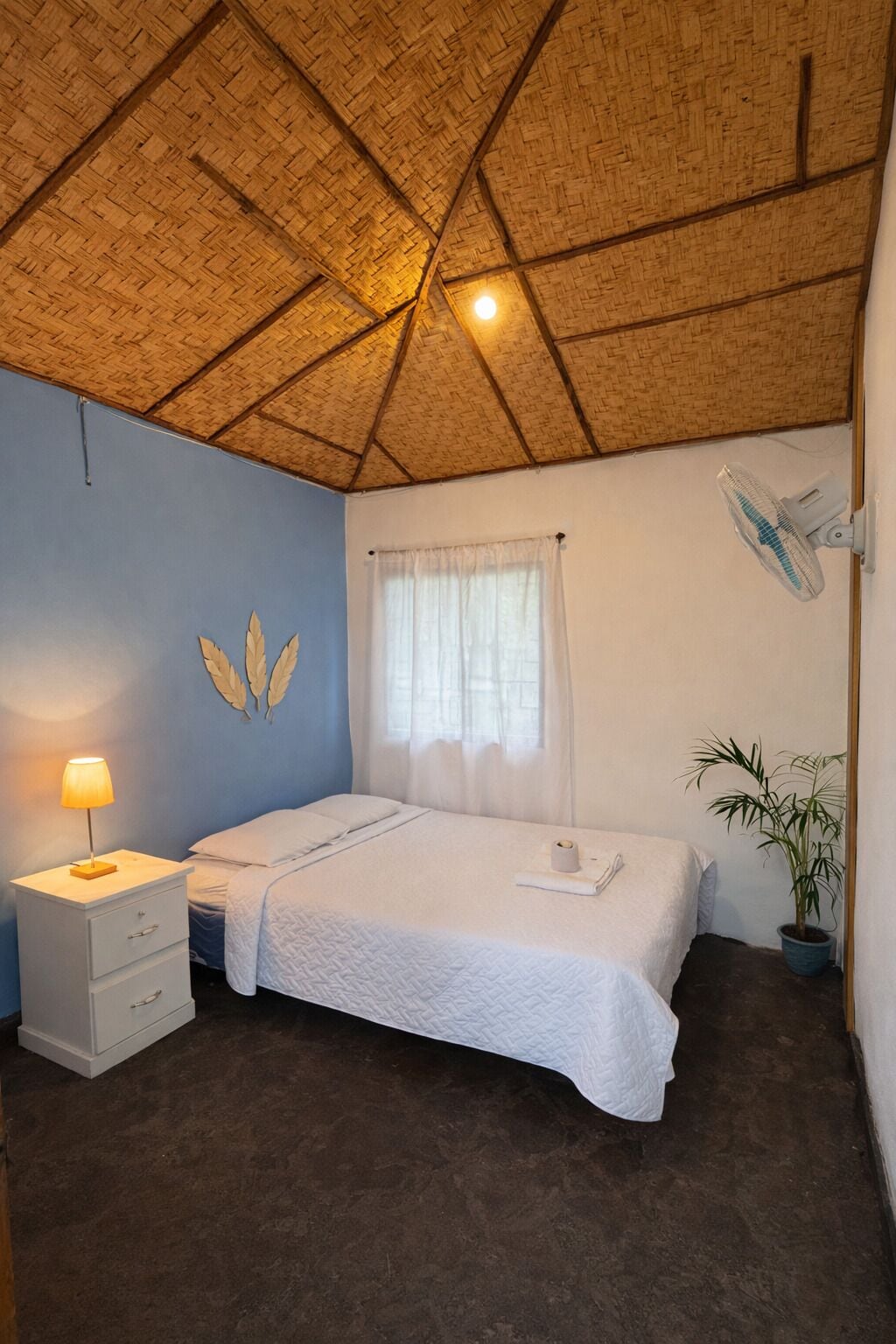 Ein Zimmer mit Strohdach, einem Bett mit weißer Bettwäsche, einem Nachttisch mit Lampe, einem Fenster mit Vorhängen und einer Blumentopf-Pflanze.