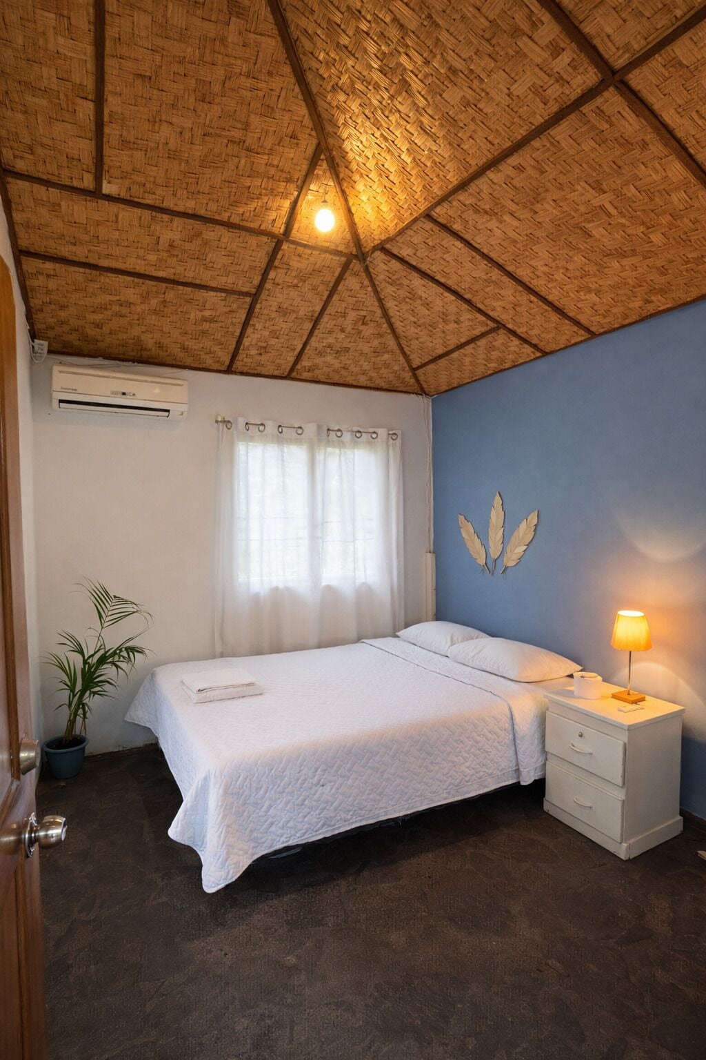 A bedroom with a thatched ceiling, a bed, a bedside table with a lamp, a potted plant, and an air conditioning unit.