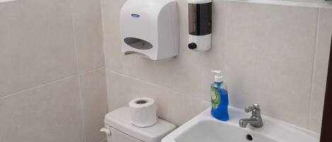 A bathroom with a toilet, a sink, a trash bin, a soap dispenser, and a toilet paper roll.