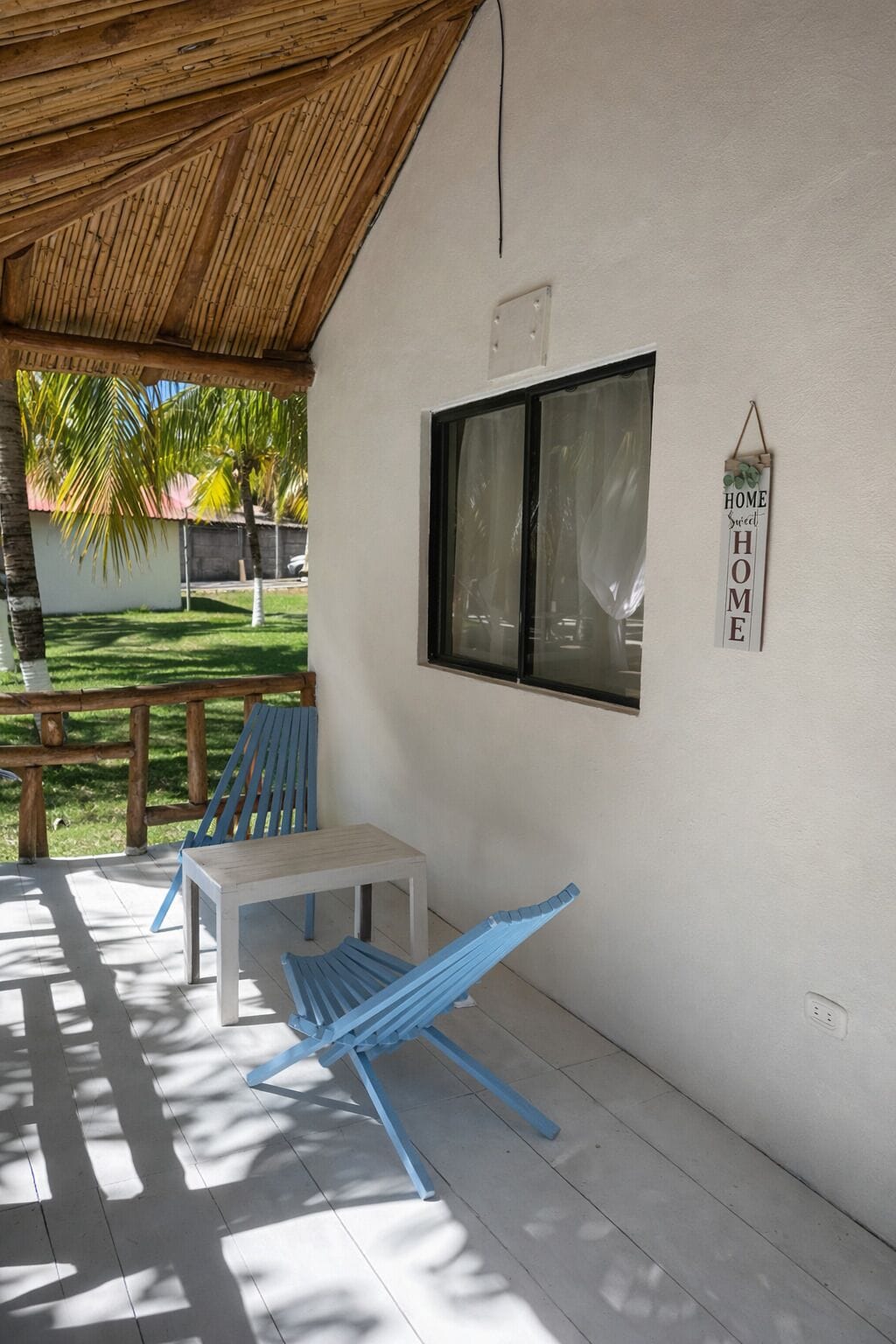 A covered patio with a table and blue chairs, a window with a 'HOME Sweet HOME' sign, and a view of a green lawn and palm trees.
