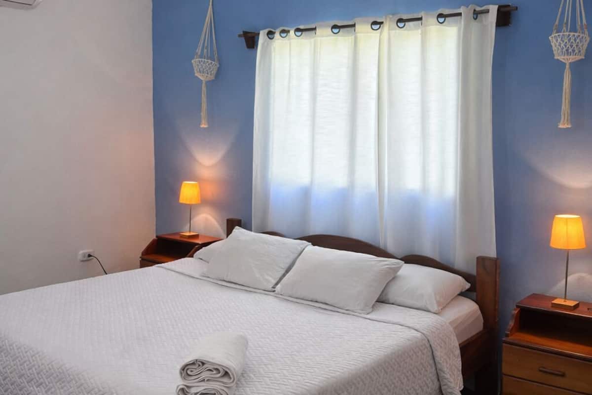 Ein ordentlich bezogenes Bett mit weißer Steppdecke, zwei Nachttischlampen und einer hängenden Lampenschirm aus Makramee.