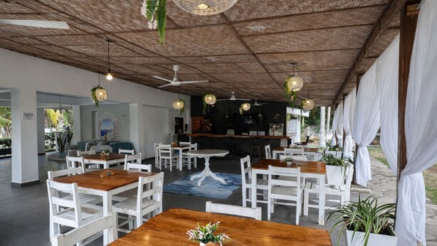 A restaurant with wooden tables and white chairs, a bar area, and hanging lights.