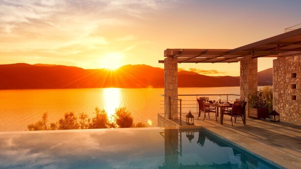 An infinity pool with a view of a sunset over a body of water, a stone structure, and outdoor furniture. - Stylish Contemporary Villa with Sea Views and Steps to the Water (Meganissi)