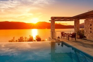 Une piscine à débordement offrant une vue sur un coucher de soleil se reflétant sur une étendue d’eau, ainsi qu’une structure en pierre et du mobilier d’extérieur.