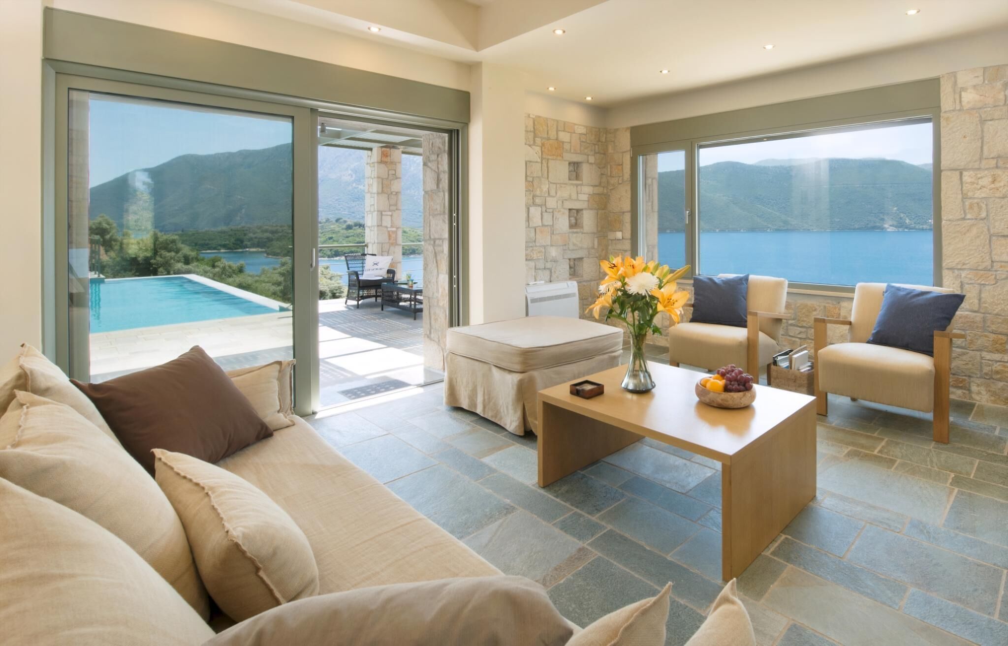 A modern living room with a large sofa, a coffee table with a vase of flowers, and a view of a pool and mountains through large windows.
