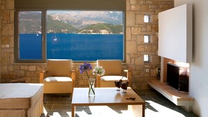 A room with a stone wall, a large window with a view of the sea and mountains, two chairs, a table with a vase of flowers, and a flat-screen TV. - Stylish Contemporary Villa with Sea Views and Steps to the Water (Meganissi)