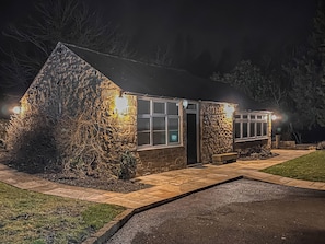 A stone building with a glass door and windows, illuminated by exterior lights. - Church Cottage nestles in the grounds of the Old Vicarage in Darley Bridge. (Darley Bridge, near Matlock)