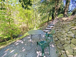 A garden patio with a round table and two chairs, surrounded by a stone wall and a gravel path.