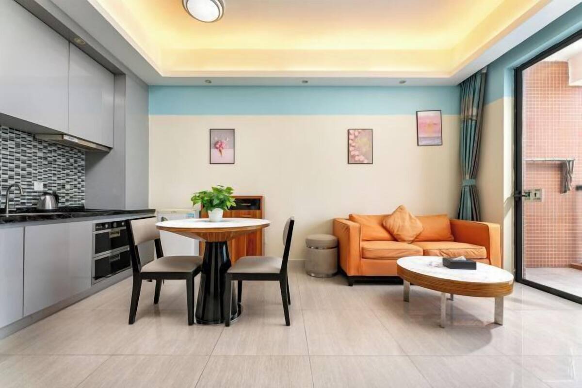 Une cuisine moderne avec des armoires grises, un comptoir noir et une hotte en acier inoxydable. On y trouve un coin repas avec une table ronde, deux chaises et une plante en pot. Un canapé orange est placé contre le mur, et une petite table ronde avec un livre est posée sur le sol.