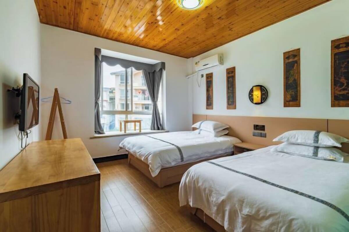 A room with two beds, a wooden desk, a window with curtains, and wall-mounted artwork.