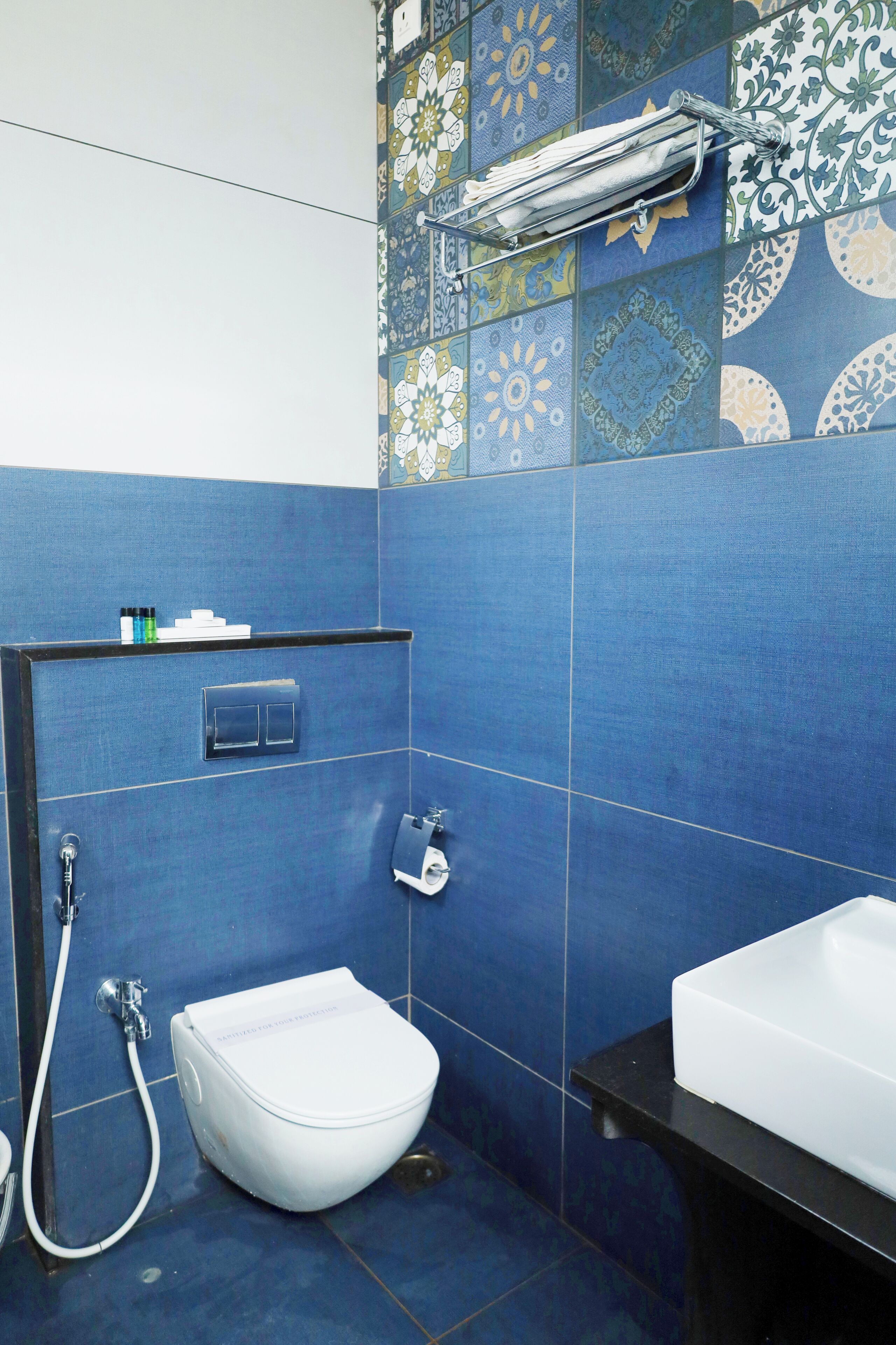Un baño con azulejos azules, un inodoro blanco, un lavabo, un perchero para toallas y un cabezal de ducha.
