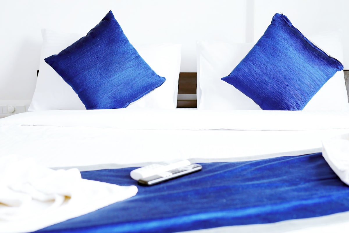 A bed with two blue pillows, a white blanket, and a remote control on the bed.
