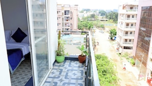 Un balcón con una cama, dos plantas en macetas y vistas a una zona residencial con edificios y vegetación.