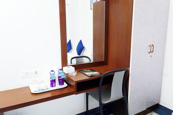 Habitación de hotel con un escritorio de madera, una silla, un espejo y una cama con almohadas azules. Hay artículos de aseo en el escritorio.