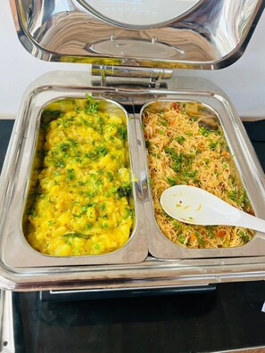 Un recipiente de acero inoxidable con dos compartimentos, uno con un plato amarillo y el otro con un plato de fideos, y una cuchara blanca en el compartimento de fideos.