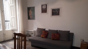Un salon avec un canapé gris, deux coussins à motifs, une chaise en bois, une petite table d’appoint blanche et une bibliothèque.