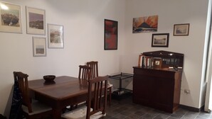 Un coin repas avec une table et des chaises en bois, une petite table d’appoint avec un bol, et une bibliothèque remplie de livres.