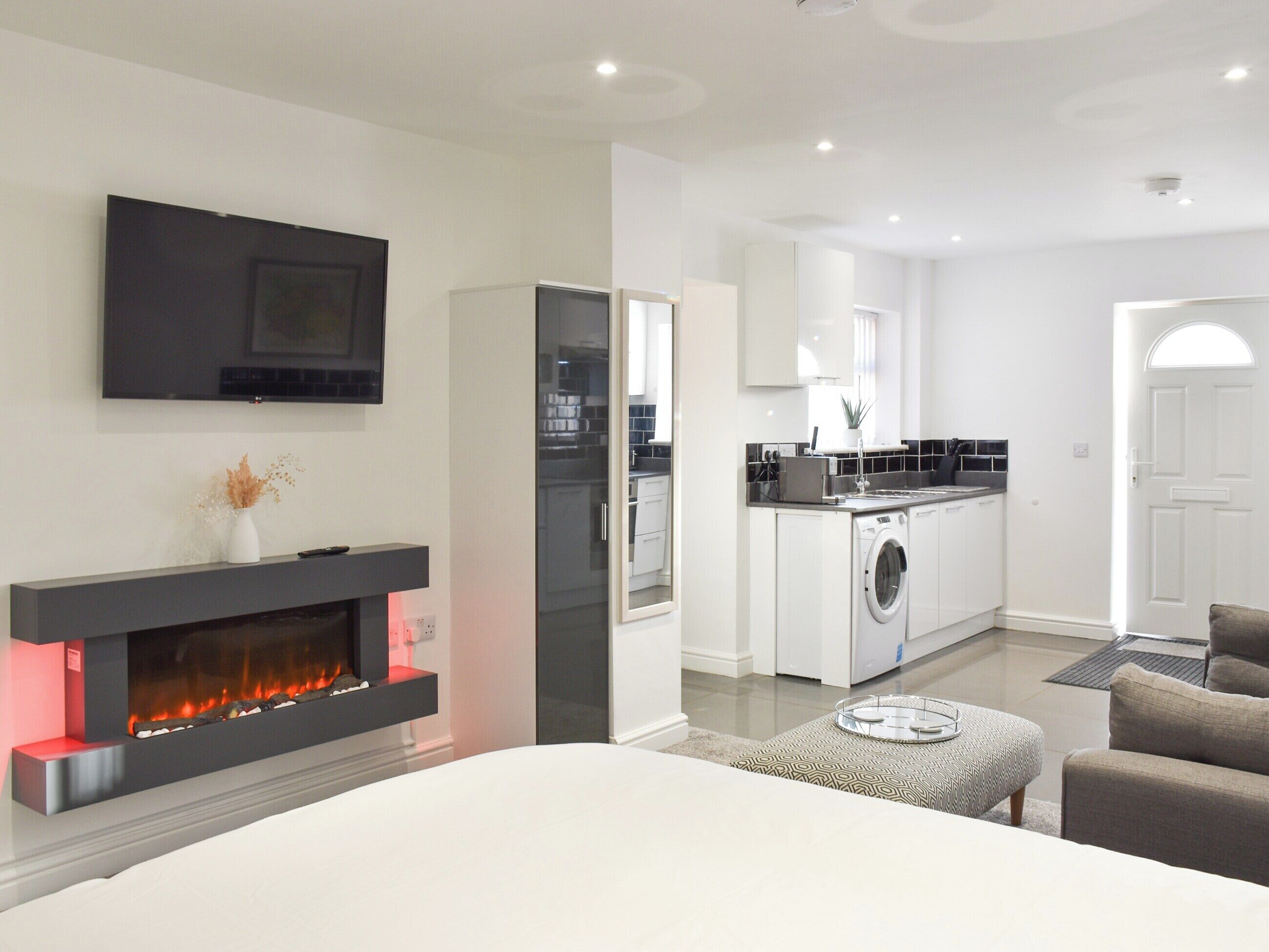 A modern living room with a fireplace, a TV mounted on the wall, and a kitchen area with a washing machine.