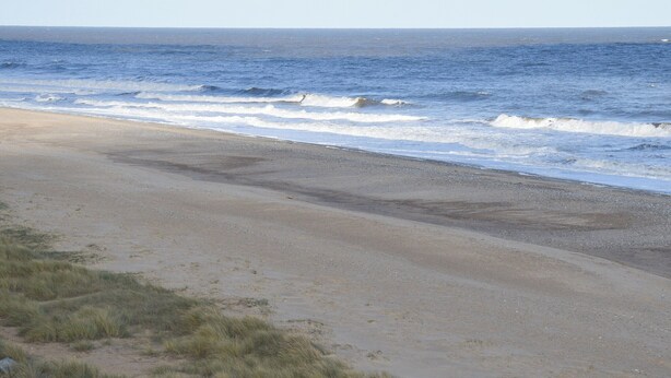 Een strand met golven die de kust naderen, een heldere lucht en land in de verte.