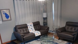 A living room with a brown leather sofa, a glass coffee table, a blue and white patterned rug, a wooden floor, and a ceiling fan.