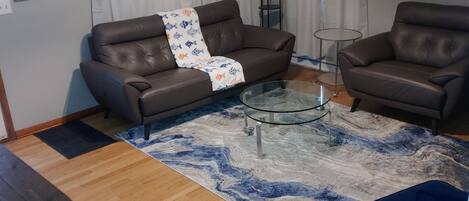 A living room with a brown leather sofa, a glass coffee table, a blue and white patterned rug, a wooden floor, and a ceiling fan.