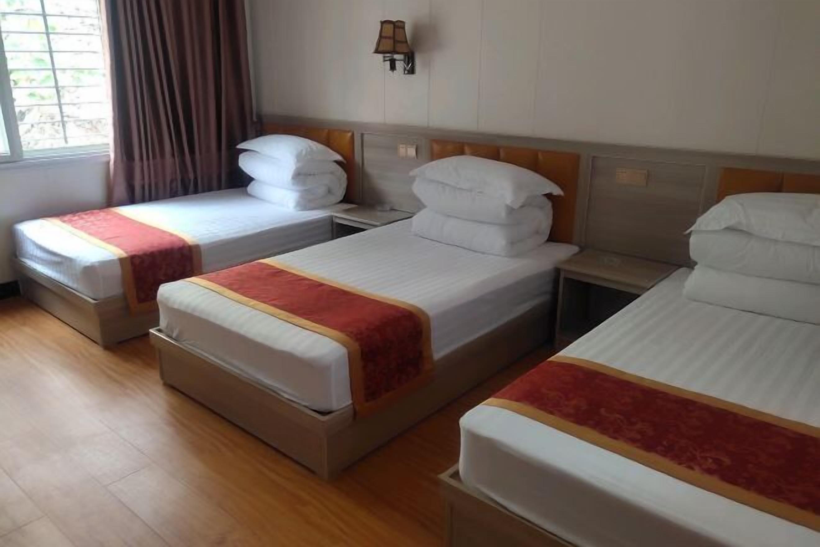 A hotel room with three single beds, each with a red and white bedspread, wooden headboards, and a nightstand with a lamp.